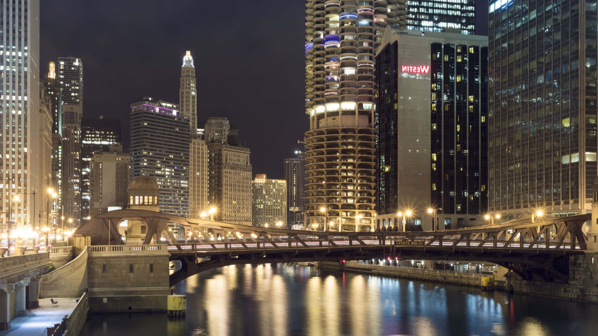 Nighttime city skyline with lit buildings and bridge over water.
