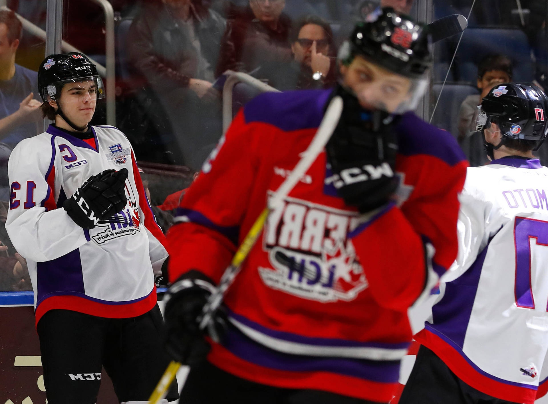 Hockey players in red white and purple jerseys on ice during game with spectators behind glass.
